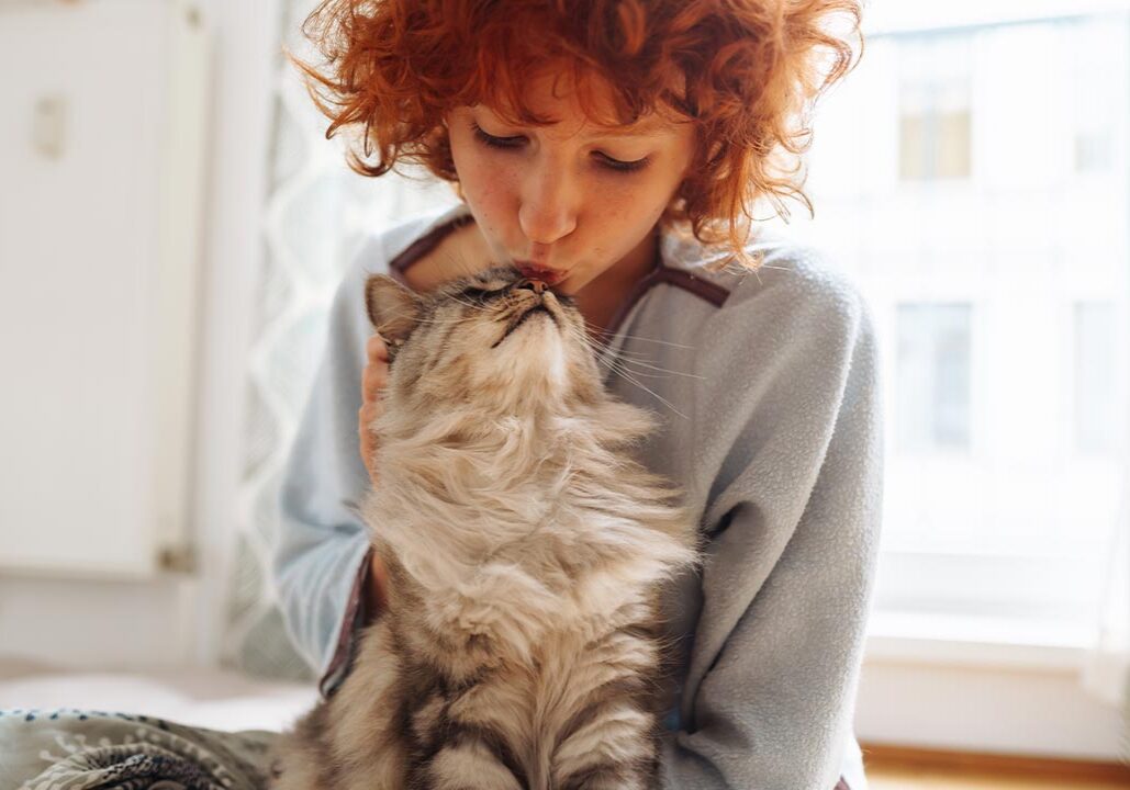 female with red curly hair kissing fluffy cat's nose while holding and petting cat indoors